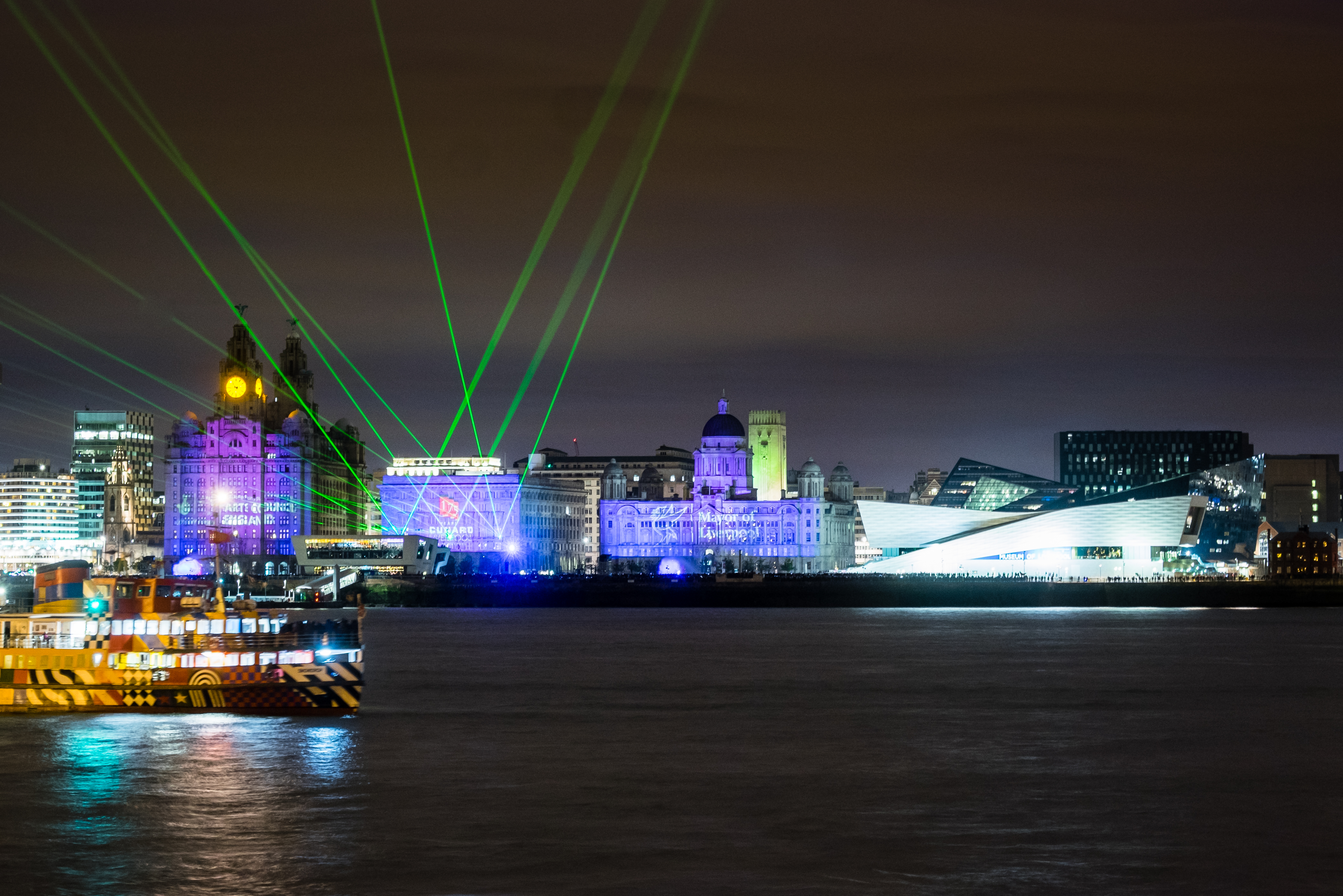 liverpool waterfront by night with the waterfront buildings lit in blue, there are green strobe lights above the cunard building and the mersey ferry is light up on the mersey - image to accompany the about culture liverpool article