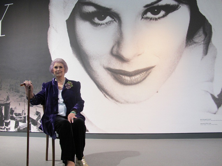 April Ashley sat on chair in front of giant photograph of her in the museum of liverpool