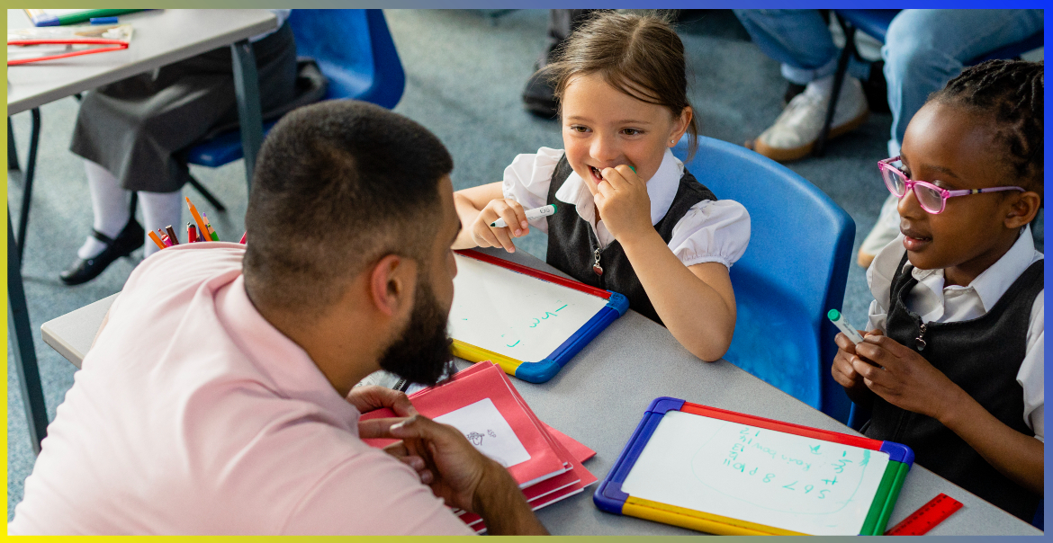 A teacher speaks to two primary school pupils at their table with whiteboards.