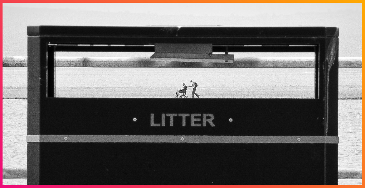 A black and white photograph of a public rubbish bin in West Kirby. Through the gap, you can see two people walking around the Marine Lake.