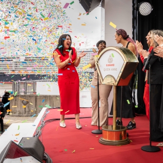 On stage for the naming ceremony of Queen Anne, five women pull the lever to name the ship. The ship and confetti can be seen behind them.