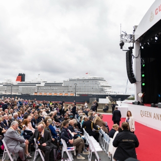 A large crowd over looking the Queen Anne cruise ship on an overcast day. A large, outdoor stage stands in front of the crowd with Cunard branding.