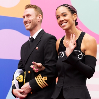 A woman and man link arms and wave at the crowd as they walk at an outdoot event.