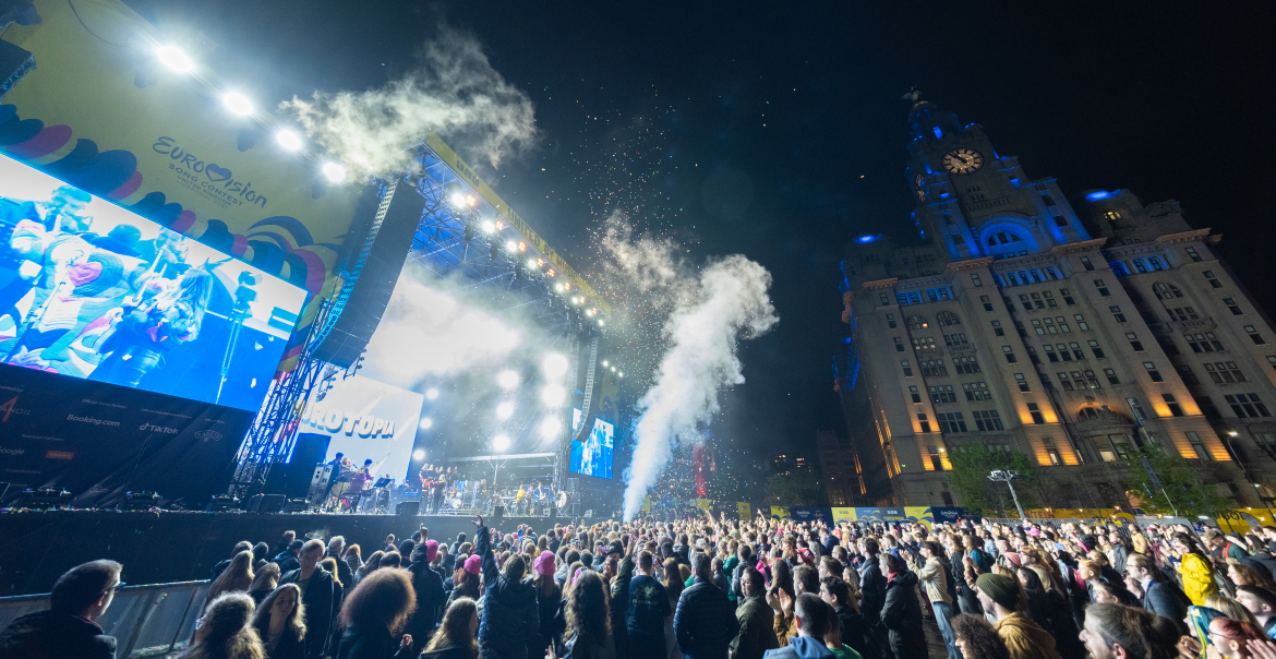 An image of a large crowd at an outdoor concert at night. The Royal Liver Building can be seen in the background.