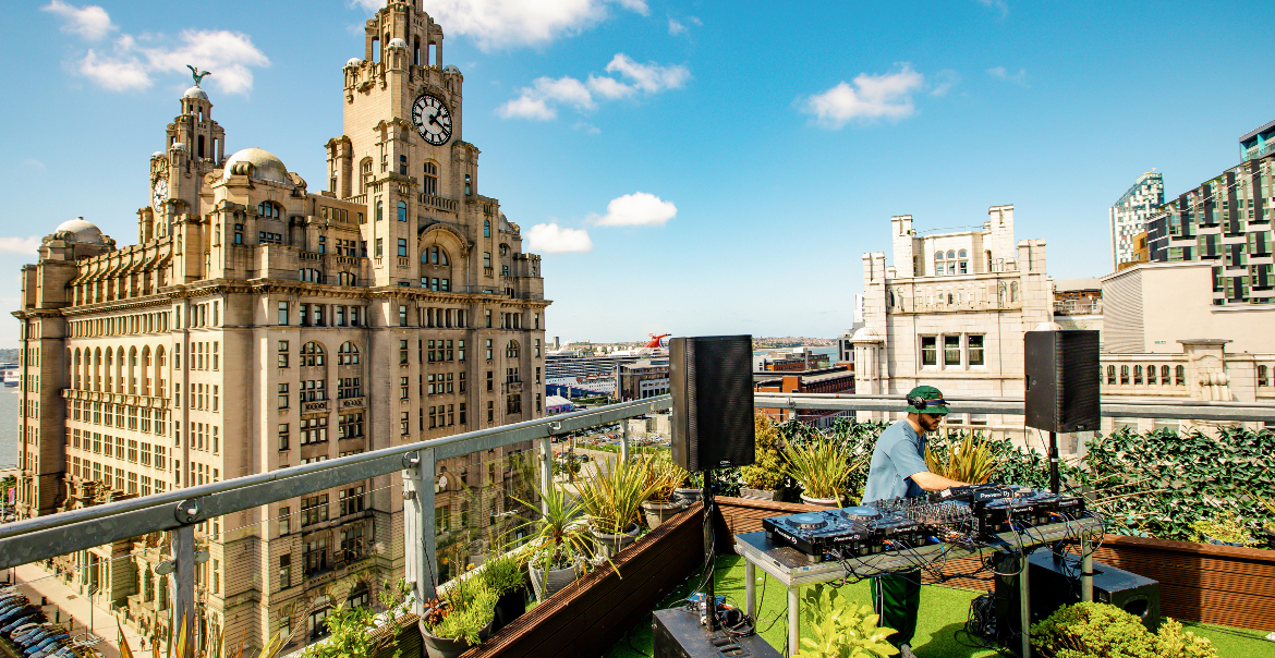 An image of a DJ performing at an rooftop bar in front of the Royal Liver Building on a sunny day