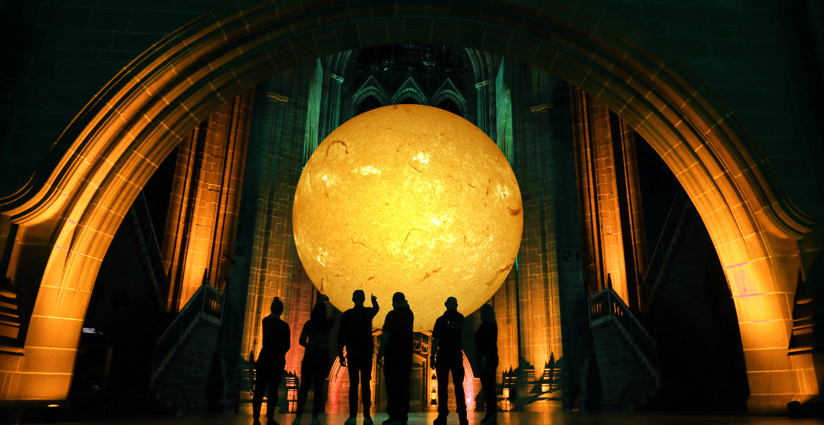 Silhouettes of several people observing a large, illuminated sun installation inside of Liverpool Cathedral.