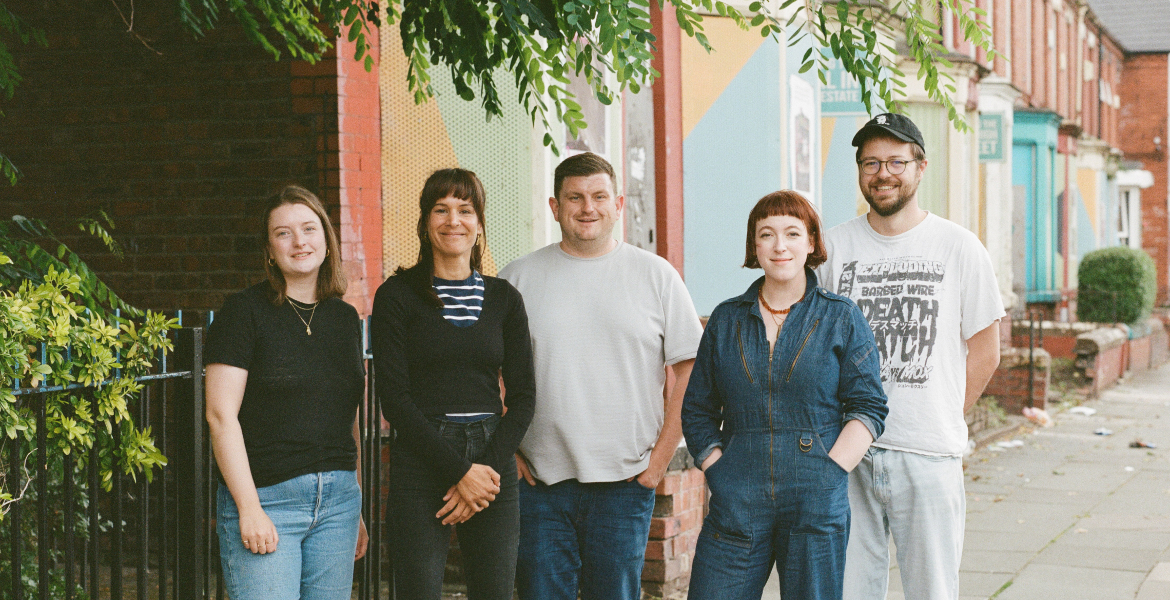 five staff members of Homebaked in Liverpool stood in front of terraced houses with tree leaves hanging above their heads