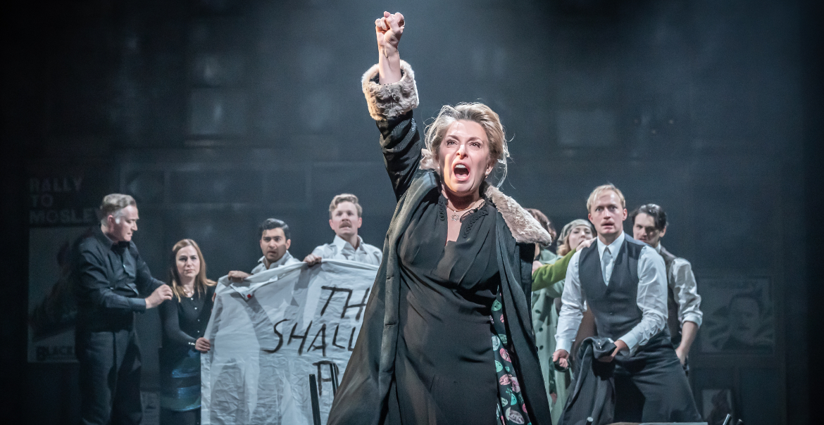 A production image of The Merchant of Venice. In this scene, a character stands with their right up up and a group behind them